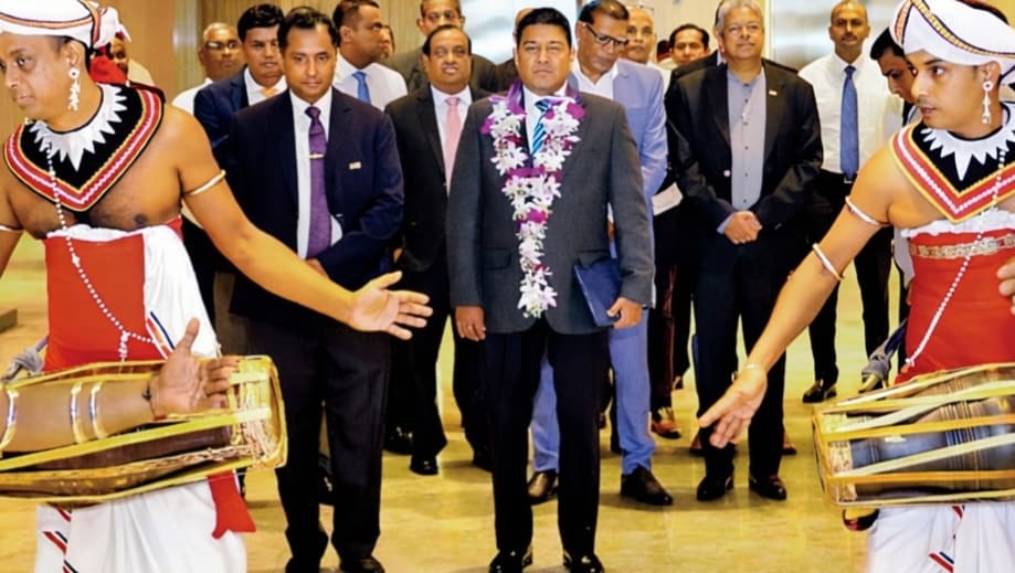 Traditional drummers perform a ceremonial welcome as a garlanded man in a suit stands centered, surrounded by guests.